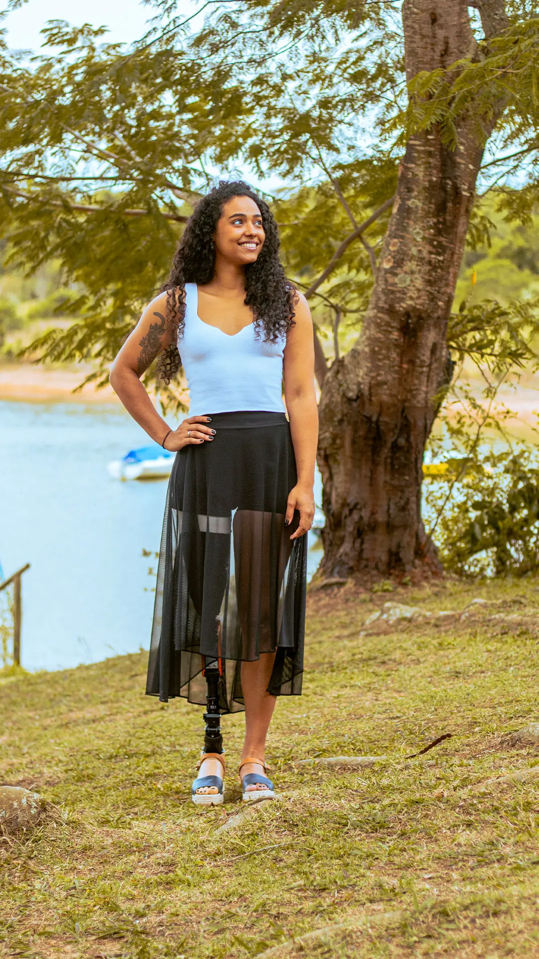 Mulher de saia com prótese de perna, de pé num bosque à beira do lago